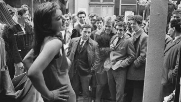Young men gather around a boutique window where a model stands with her bare breasts…